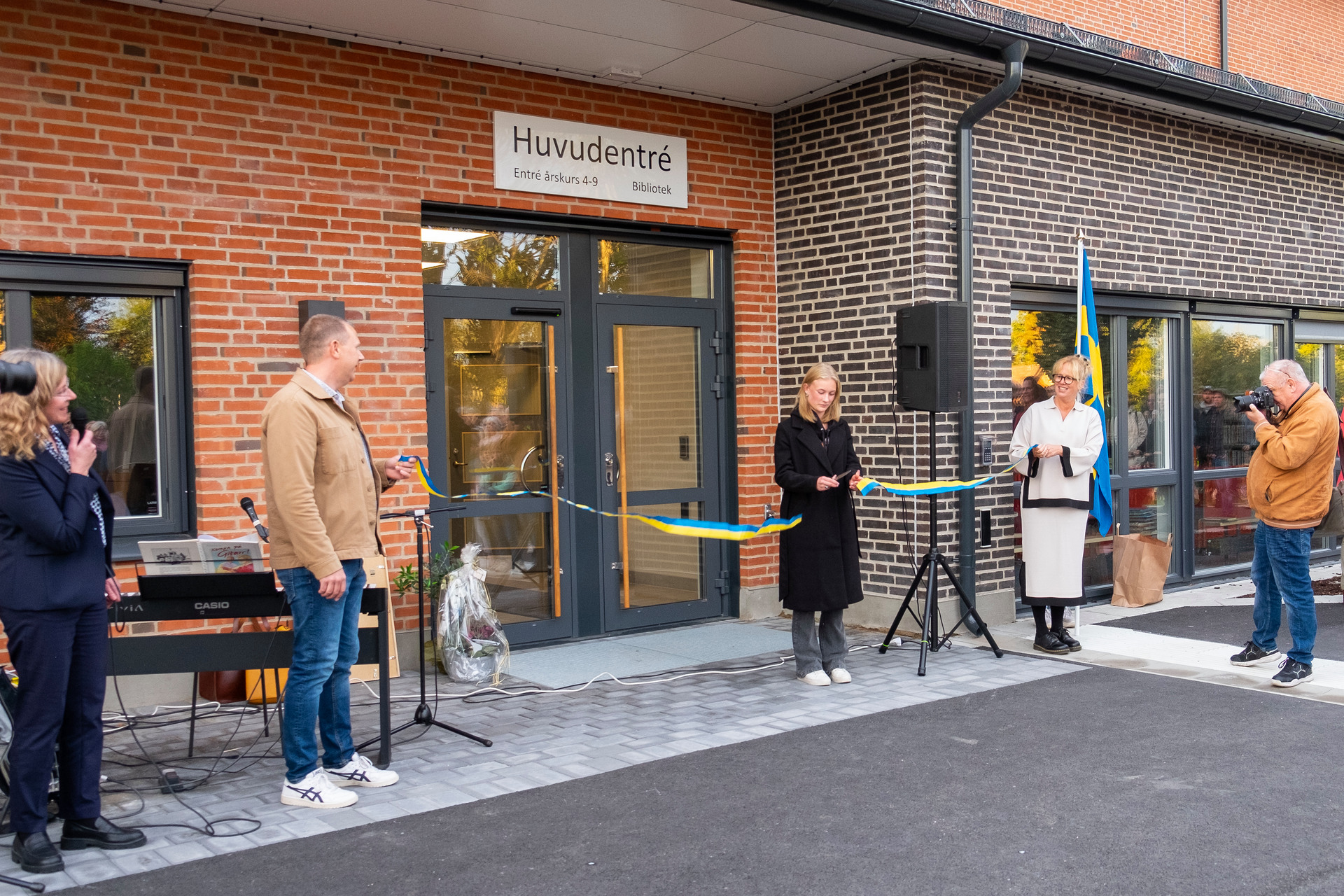 Ett blågult band är sträckt mellan två politiker. En elev i en svart rock klipper bandet. Många människor står runtomkring och tittar. I bakgrunden ser man skolans entr+e.